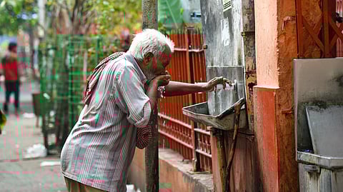 Elderly man drinks from a street water tap, leaning over and cupping water to his mouth. He wears a striped shirt, with greenery in the background.