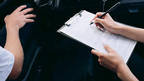 A person writes on a clipboard filled with forms, sitting in a car's passenger seat. Another person’s arm is visible nearby.