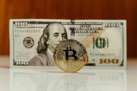 A Bitcoin coin stands upright in front of a blurred 100-dollar bill with Benjamin Franklin's portrait. The scene contrasts digital and traditional currency.