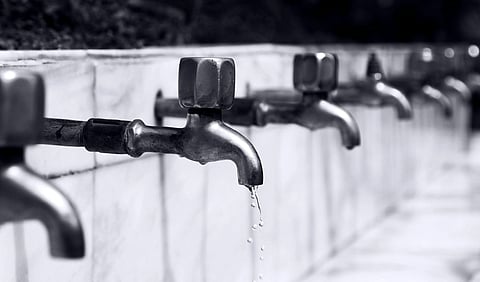Close-up of several metal water taps lined up on a white tiled wall; one tap drips water. The image conveys a mood of scarcity and conservation.