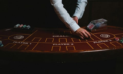 A casino dealer in a white shirt and vest deals cards at a dimly lit poker table labeled for multiple players. Stacks of chips are visible, creating a tense atmosphere.