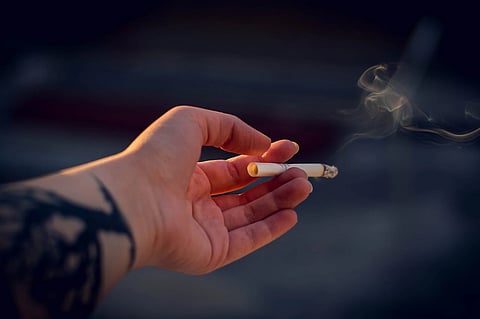 The image depicts a hand holding a lighted cigarette, with smoke coming from it.