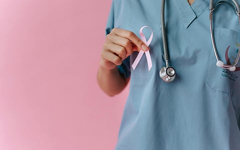 A healthcare professional in blue scrubs holds a pink ribbon and a stethoscope against a pink background, symbolizing breast cancer awareness and support.