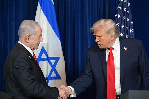 Benjamin Netanyahu (left) shakes hand with Donald Trump (right). Both leaders are wearing suit and red tie, and the flags of their respective nations are behind them.