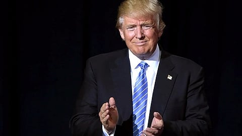 Image of US president Donald Trump set against a black background. he is wearing a dark blue suit with a light  blue tie and an American flag themed brooch. He is seen like he is making an applause.