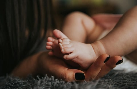 Close-up of a baby's feet gently resting on an adult's hand with black nail polish. Soft, warm lighting creates a nurturing and tender atmosphere.