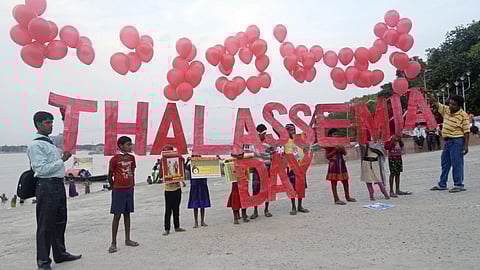 Children holding "Thalassemia Day" sign with red balloons, standing on a riverside promenade. The scene conveys awareness and hope.