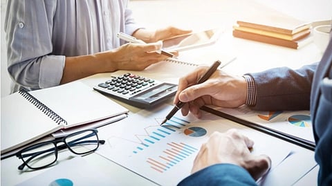 Two people in a meeting review financial charts and graphs. A calculator, tablet, and notebooks are on the table, suggesting a focused business discussion.