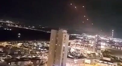 Nighttime cityscape featuring bright city lights, tall buildings, and a dark sky with glowing lights possibly indicating an aerial phenomenon.