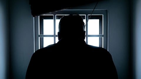 A silhouetted figure stands facing a bright window with bars, casting a mood of solitude and contemplation in a dimly lit room.