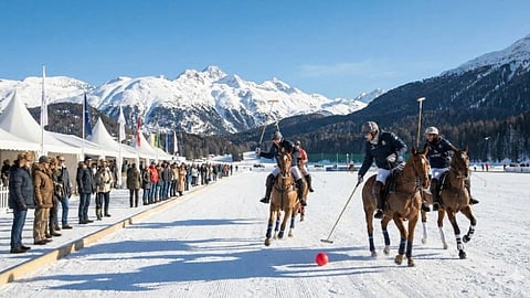 Three polo players on horseback compete on snow, chasing a red ball. Spectators stand by white tents. Snowy mountains and a clear blue sky form the backdrop.