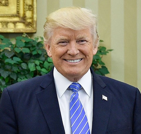 Image of Trump smiling. he is wearing his dark blue suit with a light blue stripped tie along with a brooch of the american flag. the background has beige wall paint and green plant. 