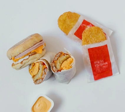 Breakfast items on a white surface: a ham and cheese muffin, two wrapped hash browns, a burrito cut in half, and a small cup of sauce.