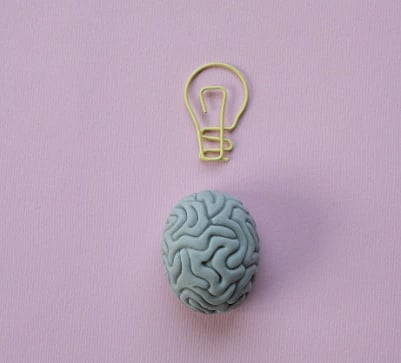 A small model of a gray brain sits on a pink background, with a yellow lightbulb paperclip above it, symbolizing ideas and creativity.
