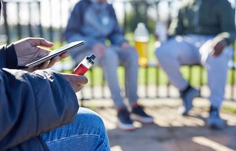 A person holding a vape and smartphone sits on a bench, with two others blurred in the background. Casual atmosphere, sunny outdoor setting.
