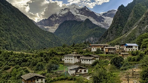 A lush, green valley with scattered traditional houses nestled against a backdrop of towering mountains partially covered by clouds and snow.