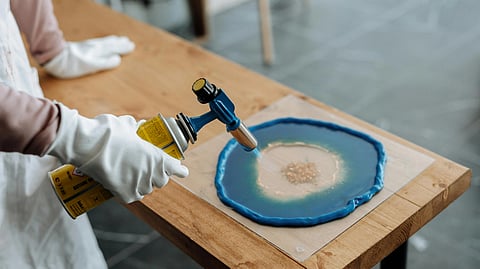 A person in gloves uses a torch to create art on a table. The artwork features blue and beige resin spread in a circular pattern.