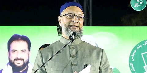 The image shows Owaisi speaking at a rally in Mumbai, he is wearing a green Sherwani and a blue cap, behind him is a poster that shows Imtiaj Jaleel.