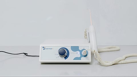 Close-up of a medical ozone therapy machine on a white surface. It has a control knob, indicator lights, and a connected handheld device with a coiled cord.