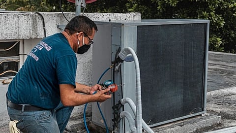 A technician wearing a mask and blue shirt checks an outdoor air conditioning unit on a rooftop. A red satellite dish is in the background. Trees are visible.