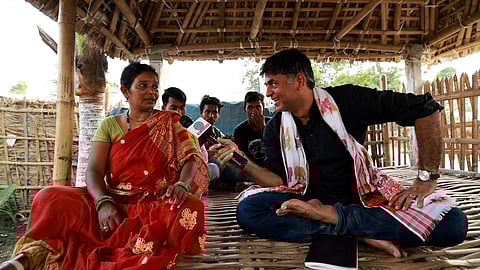 A woman in a red sari is interviewed by Saurabh Dwivedi holding a microphone in a rustic setting. Several people sit in the background, adding a community feel.