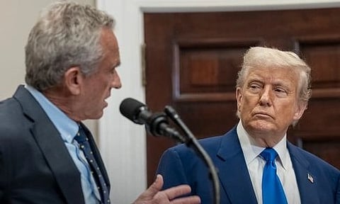 A man in a suit speaks into a microphone, gesturing, while US President Donald Trump with light hair watches attentively. They are indoors, with a wooden door behind them.
