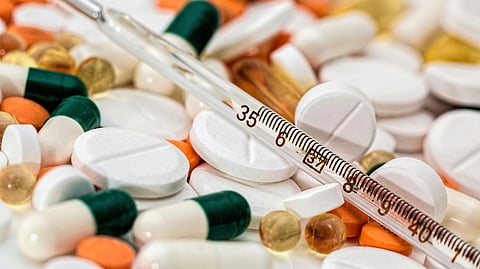 A pile of assorted pills, capsules, and gel tablets in various colors with a glass thermometer placed diagonally across them, conveying a medical theme.