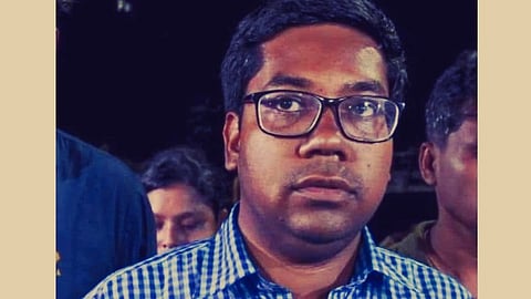 Aniket wearing glasses and a blue checkered shirt looks directly at the camera. He is in a crowd, appearing serious against a dark background.