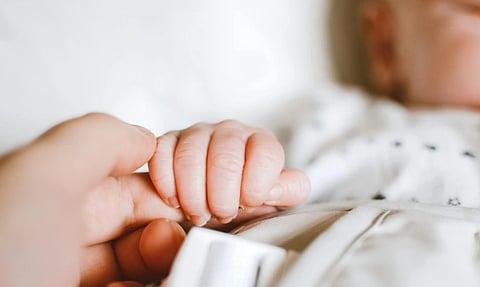 Close-up of a baby's hand gently gripping an adult's finger. The scene conveys warmth and tenderness, set against a soft, neutral background.