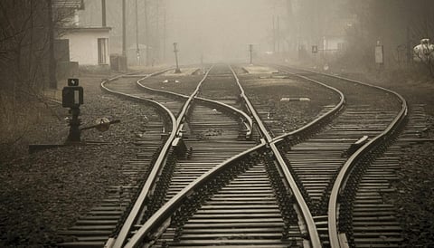 Foggy railway tracks diverge amidst bare trees and a small building, creating a mysterious, tranquil scene with muted, earthy tones.