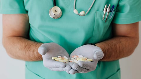 A person in green medical scrubs and gloves holds various pills in cupped hands. A stethoscope and pens are visible, conveying a healthcare setting.