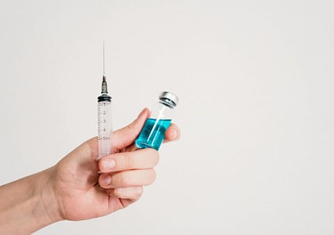 A hand holds a syringe and a vial filled with blue liquid against a plain white background. The image conveys themes of medicine and healthcare.