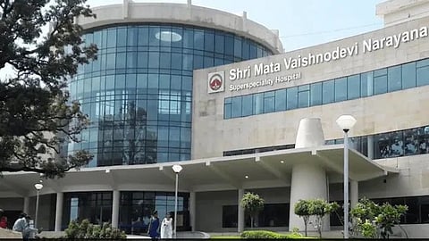 Modern hospital exterior with a large glass facade and a sign reading "Shri Mata Vaishnodevi Narayana Superspeciality Hospital." People and trees in foreground.