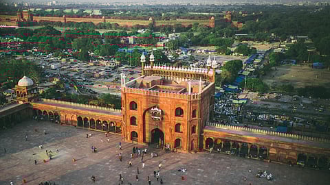 Aerial view of Jama Masjid in Delhi, showcasing red sandstone architecture. The expansive courtyard is dotted with people, exuding a lively, bustling atmosphere.
