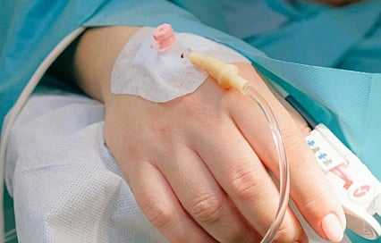 A close-up of a hand with a hospital IV catheter inserted, resting on a bed. The person wears a teal medical gown, evoking a sense of hospital care.