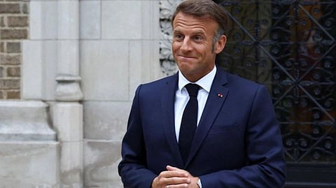 French President Emmanuel Macron in a suit stands outside with a lighthearted smile and clasped hands. The background features ornate stone architecture and a decorative metal gate.