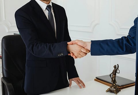 Two men in suits are shaking hands in an office setting, symbolizing agreement or partnership. A Lady Justice figurine is on the desk beside them.
