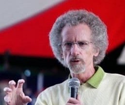 Image of Christian Author Philip Yancey speaking at the Greenbelt Festival at Cheltenham Racecourse in England on August 24, 2008. he speaks into a microphone, gesturing with his hand. He wears a light sweater, standing against a blurred red and white background.