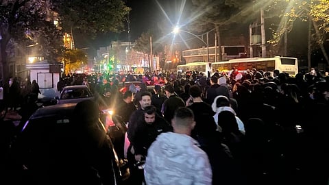 A large crowd fills the street of Iran at night, illuminated by streetlights and vehicle headlights.