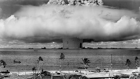 A powerful black-and-white image shows a massive mushroom cloud rising from a nuclear explosion over the ocean, palm trees in the foreground, evoking a tense atmosphere.