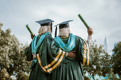 Two graduates in green caps and gowns, draped in blue sashes, embrace while holding diplomas, celebrating under a cloudy sky amidst greenery.