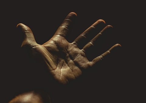A close-up of an open hand reaching out in darkness, lit by soft light. The detailed texture of the palm adds an intimate, hopeful tone.