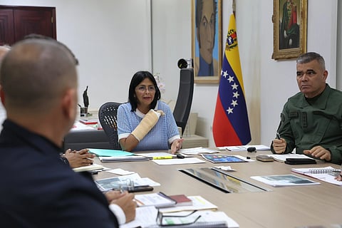 Delcy Rodriguez in her cabin, surrounded by government officials. Venezuelan flag is to her left, and documents lying on the table.