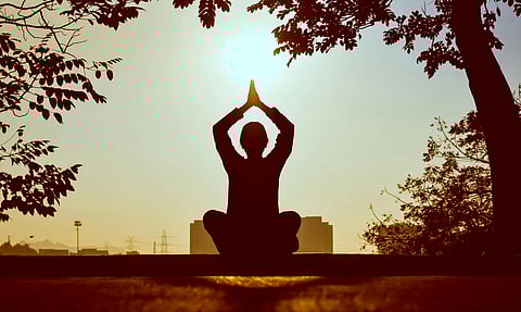 A person meditates in a cross-legged position with hands in prayer above their head, silhouetted against a sunrise, surrounded by tree branches. Calm and peaceful mood.