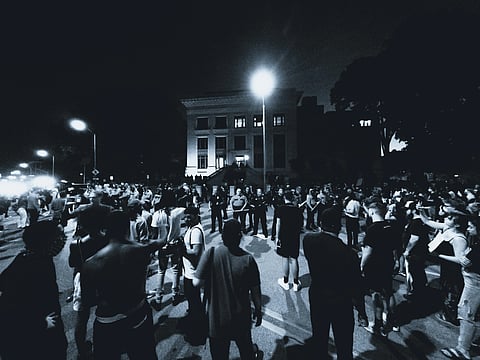 Large crowd gathers at night outside a well-lit building, facing a line of officials or security personnel. The atmosphere appears tense and serious.