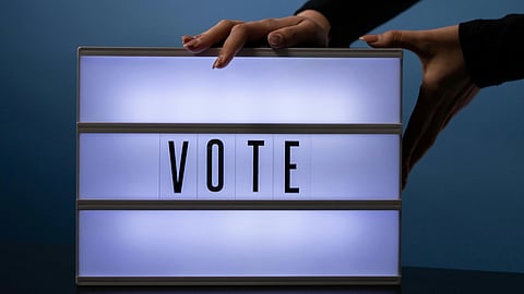 Hands adjust a glowing lightbox displaying the word "VOTE" against a blue background, conveying a theme of participation and action.