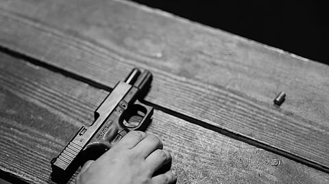 A hand reaches for a black pistol on a wooden table, with a single bullet nearby. The scene is dimly lit, creating a tense and serious atmosphere.