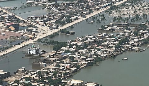 Aerial view of a flooded town with partially submerged buildings and roads. A mosque stands prominently above water, surrounded by scattered greenery. The scene conveys a sense of calm amidst devastation.