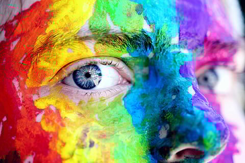 Close-up of a person's face covered in vibrant multicolored paint, highlighting a blue eye. The image conveys creativity and artistic expression.