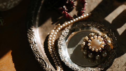 A vintage silver tray on a wooden table holds a pearl necklace, a beaded bracelet, and a pearl brooch. Warm sunlight casts soft shadows, creating an elegant, nostalgic mood.